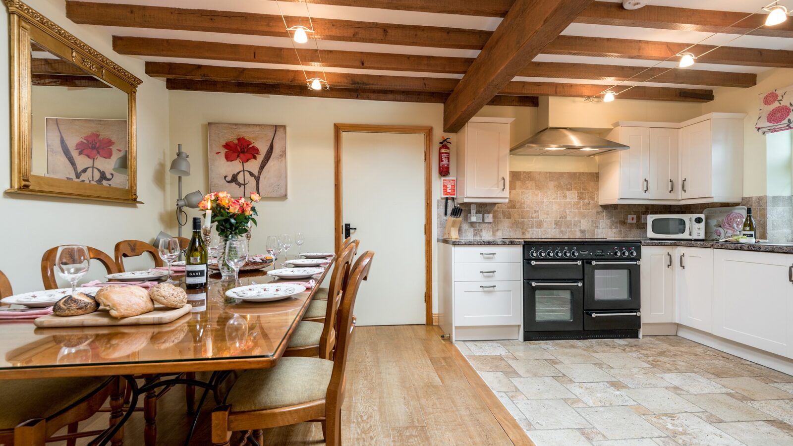 A rustic kitchen and dining area at Broadgate Farm Cottages features a wooden table set for a meal with six chairs, a bouquet of flowers as a centerpiece, and a fully equipped kitchen with white cabinets, black stove, microwave, and open shelves. Exposed wooden beams and wall art add charm.