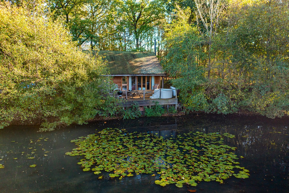 A rustic cottage with a deck, managed by Browning Bros, sits surrounded by trees near a pond with lily pads.