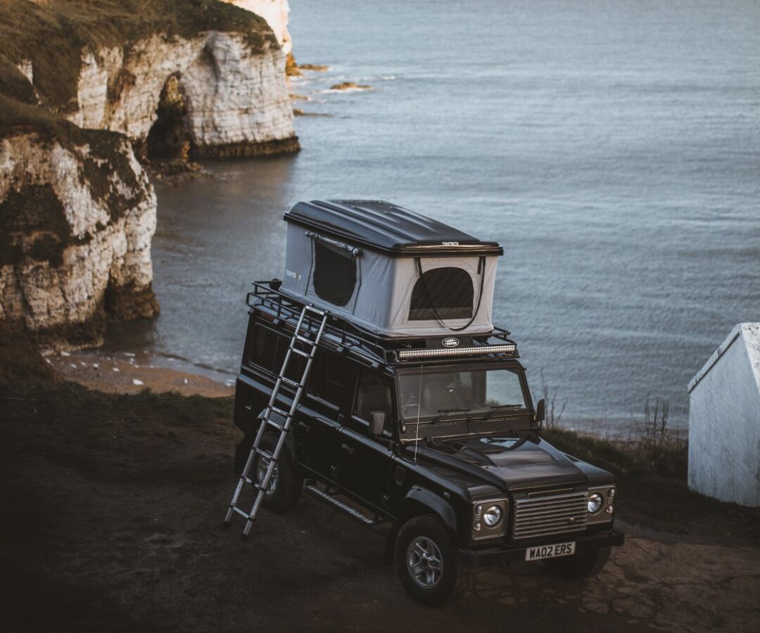 A Defender with a rooftop tent is parked by the coastal cliffs, offering the perfect camping experience as it overlooks the ocean under a clear sky.