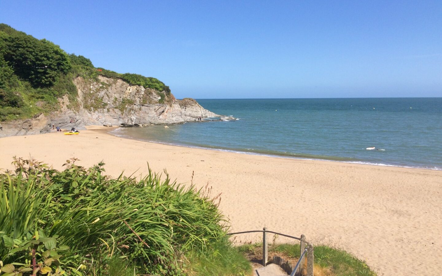 Nestled near Sloeberry Farm, the sandy beach offers serene steps leading down, surrounded by lush green cliffs under a clear blue sky.