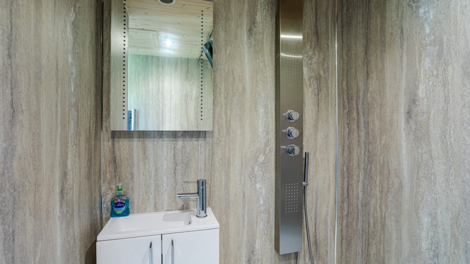 Modern bathroom with a wall-mounted mirror, a small sink with a cabinet, and a vertical shower panel on a stone-patterned wall, evoking the serene elegance of riverside cabins.