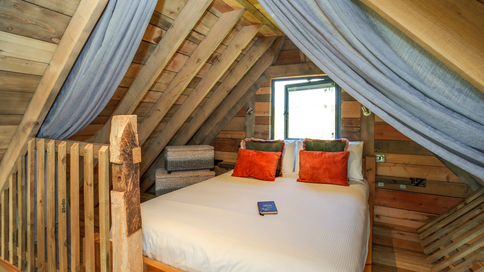 Cozy loft bedroom reminiscent of the Cadwollen Treehouse with wood-paneled walls and an A-frame ceiling. The room features a large bed with white linens, orange and green cushions, a small window, and a book on the bed, exuding rustic charm through natural decor and draped fabric accents.