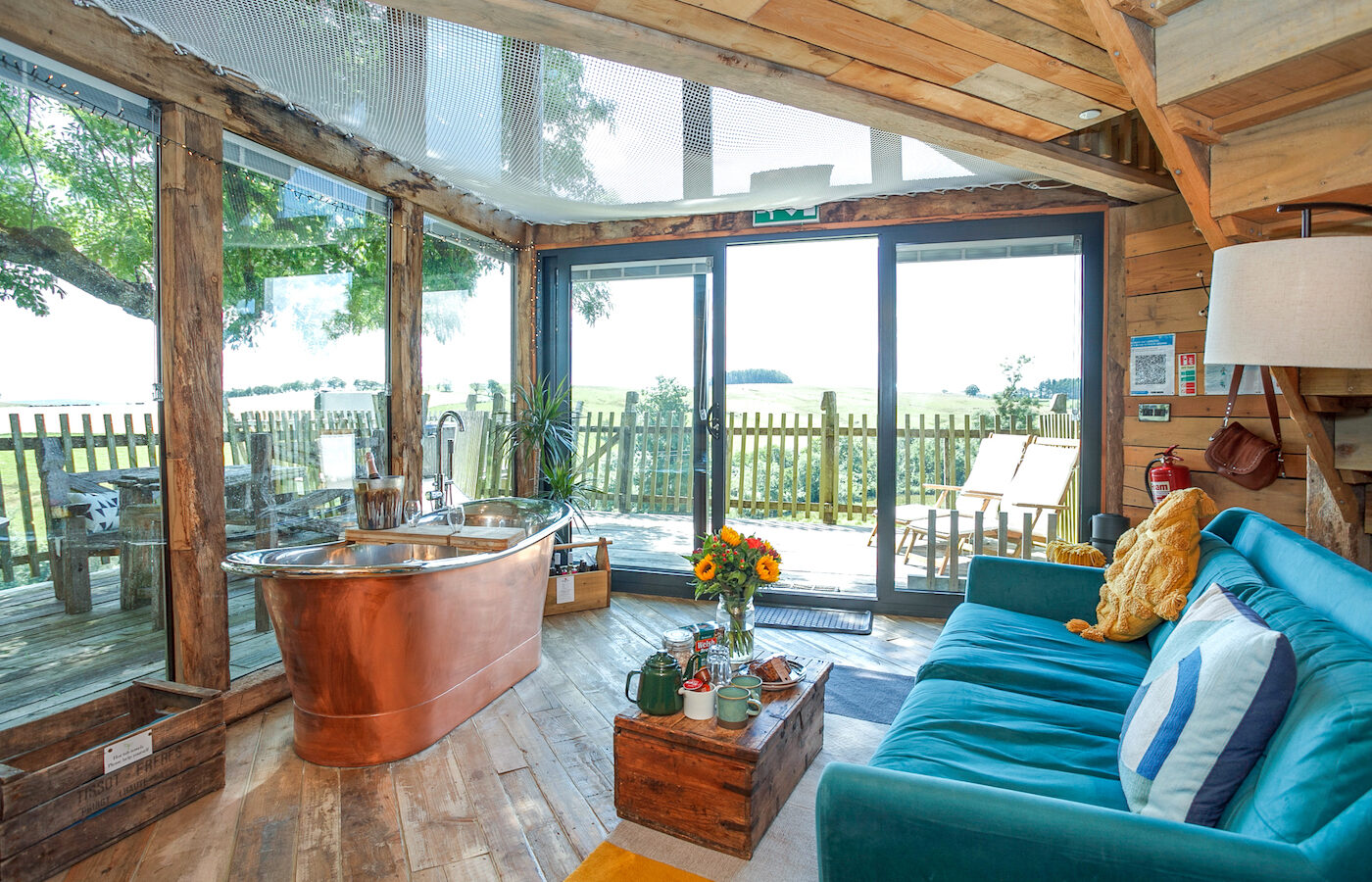 Nestled in the Cadwollen Treehouse, this cozy, rustic room boasts wooden floors and walls, a teal sofa, and a copper bathtub. A wooden coffee table adorned with flowers sits by large windows that frame a scenic view of a deck and serene countryside under the clear sky.