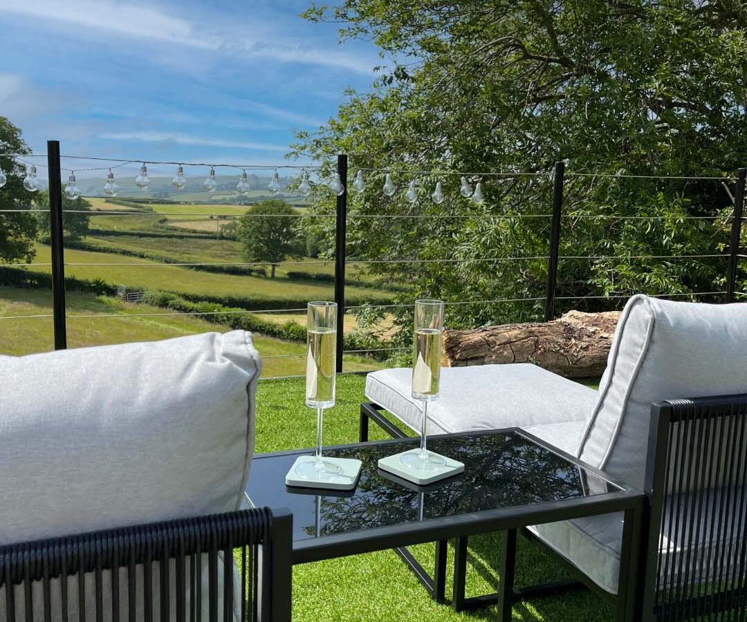 Two outdoor chairs with cushions sit on green artificial grass at Cae Cnwc Cabin, facing a scenic countryside view. A glass table between them holds two champagne flutes. String lights hang overhead, and a tree and expansive fields are visible under a blue sky.