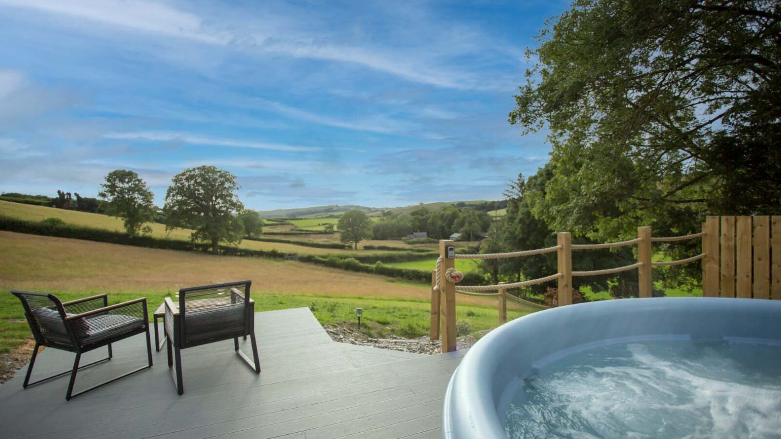 A tranquil outdoor scene unfolds at Cae Cnwc Cabin, featuring a bubbling hot tub on a wooden deck overlooking rolling green fields and trees. Two black chairs and a small table sit invitingly on the deck, creating a peaceful setting under the bright blue sky with wispy clouds.