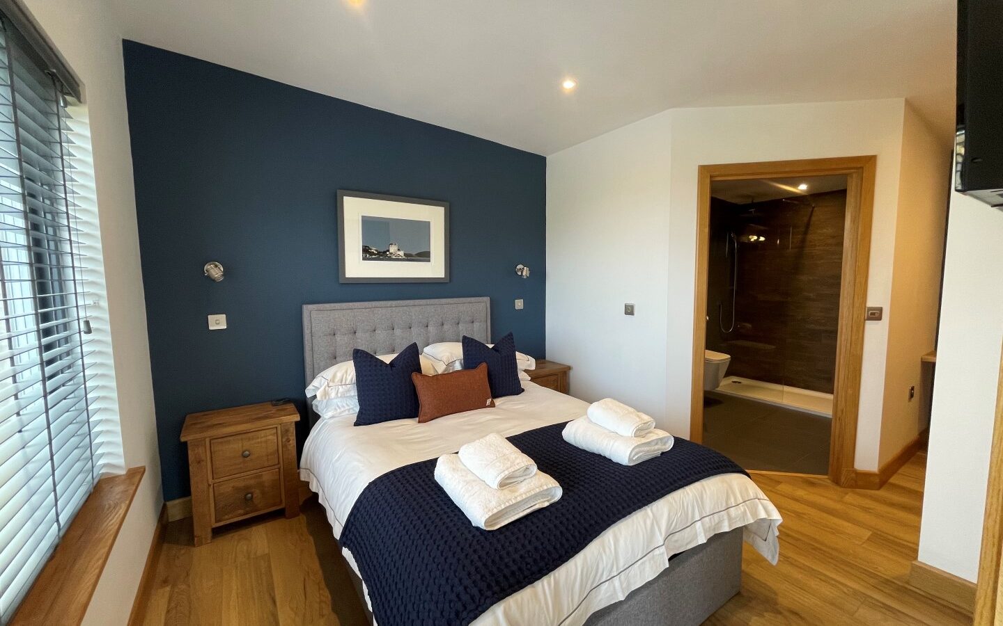 A cozy Cairn Lodges bedroom featuring a neatly made bed with white and navy bedding. Two wooden nightstands, a framed artwork above the bed, and an open doorway leading to a modern bathroom enhance the ambiance. Wooden floors and window blinds complete the decor.