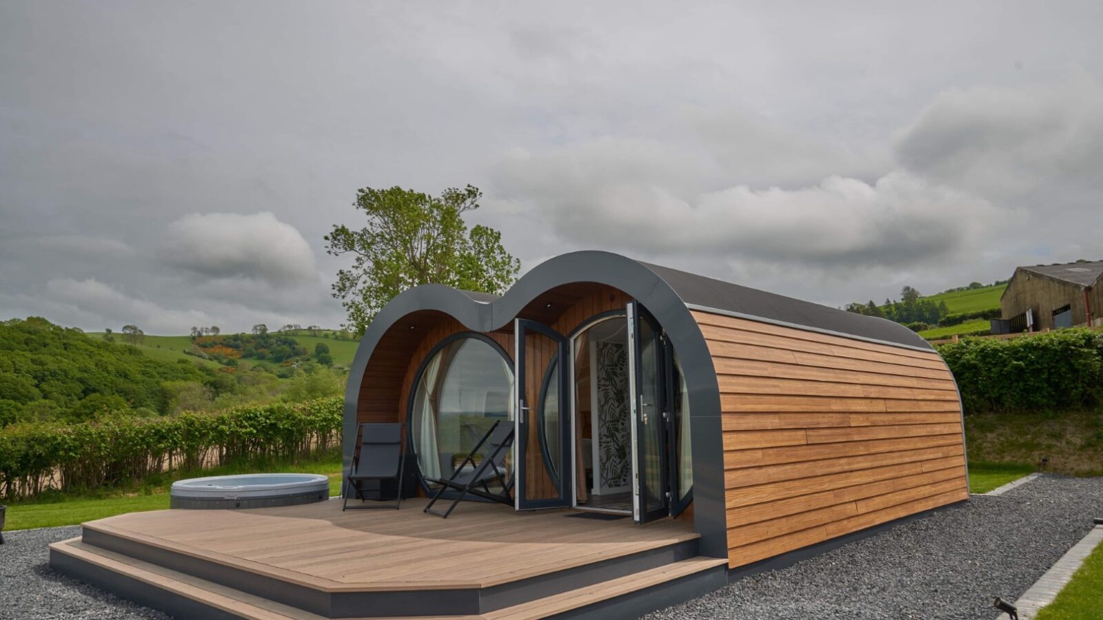 A modern, curved tiny house with wooden siding and large glass doors sits on a gravel path. This luxury pod features a deck with two chairs that offer serene views of the lush Camlad Valley's green fields and trees under a cloudy sky.