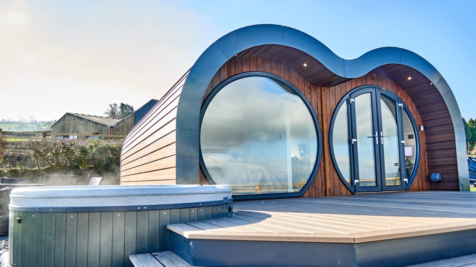 Nestled in the picturesque Camlad Valley, this luxurious, rounded wooden cabin features large circular windows and a curved roof. A hot tub graces the deck under a clear sky, offering serene views of the rural landscape—an epitome of luxury pods at their finest.