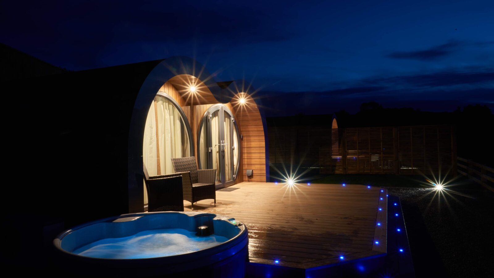 A cozy wooden cabin at night, nestled in the Camlad Valley, features an illuminated porch with a hot tub. Warm lights accentuate the curved architecture and patio area, creating a serene and inviting atmosphere reminiscent of luxury pods under a clear, dark sky.