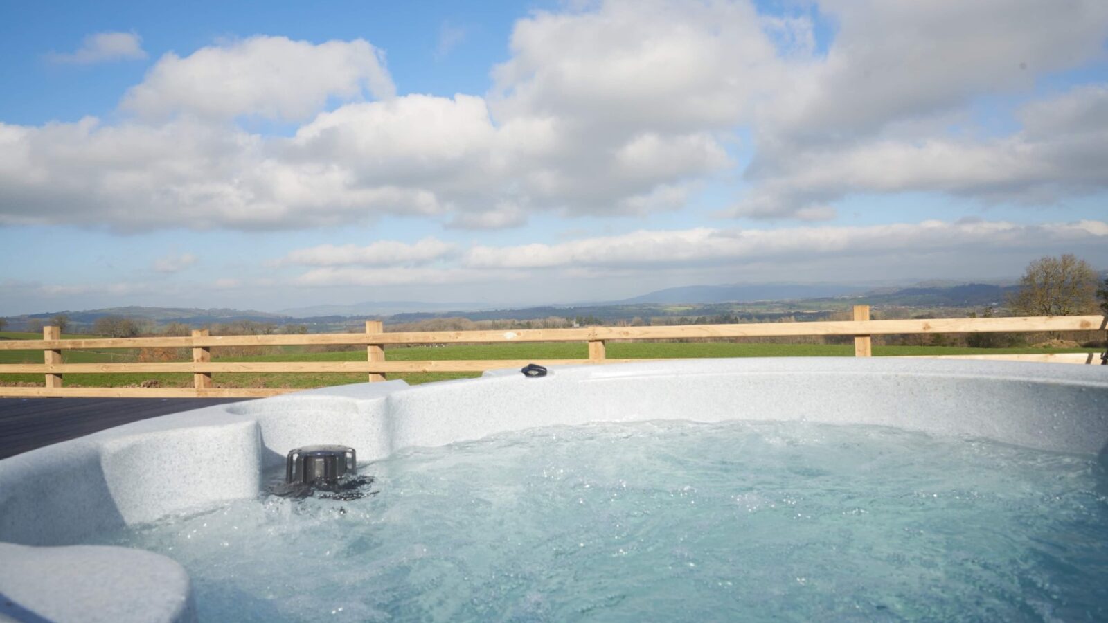 Nestled in the Camlad Valley, a bubbling outdoor hot tub offers a scenic view of green fields and distant mountains under a partly cloudy blue sky, all bordered by a wooden fence. Enjoy ultimate relaxation in this serene setting surrounded by luxury pods.