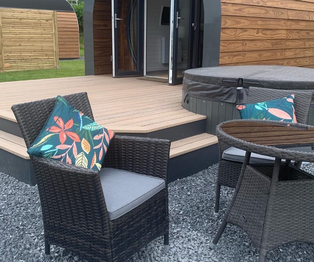 A wooden cabin with a curved roof sits on a raised deck, embodying the charm of Camlad Valley Luxury Pods. The deck features an open door and a hot tub. In the foreground, a wicker chair with colorful cushions and a small table rest on gravel, surrounded by trees and grass in the background.