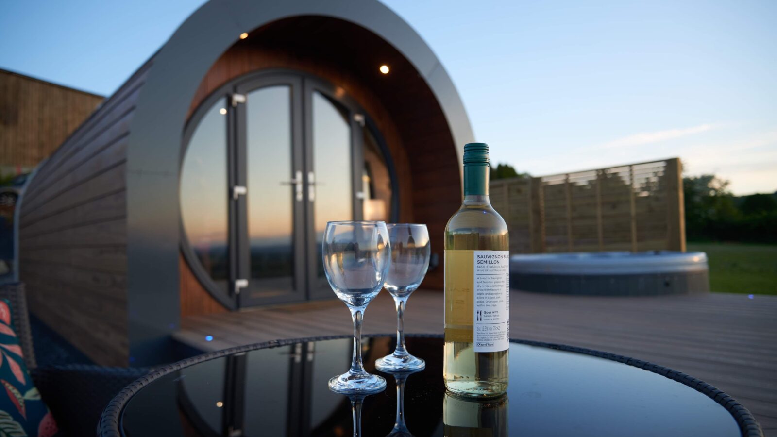 A cozy, modern cabin with a curved design is seen at sunset in the serene Camlad Valley. In the foreground, a bottle of white wine and two glasses sit on a reflective table, with a hot tub and wooden deck beside these luxury pods.