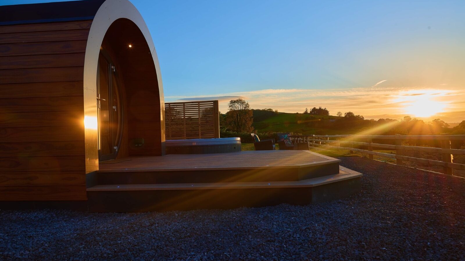 A luxury wooden cabin with a curved roof stands bathed in the sunset glow, nestled in Camlad Valley. Set on a wooden platform with stone steps, this pod features a hot tub overlooking a serene landscape, framed by clear skies and distant trees.