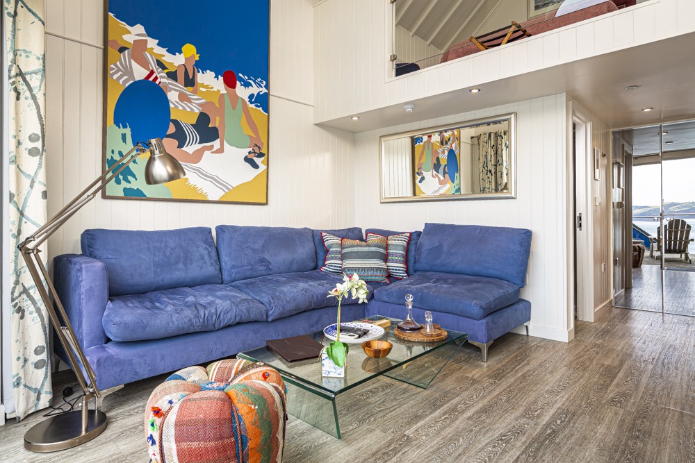 The living room features a luxurious blue sectional sofa reminiscent of the cozy suites at Cary Arms, vibrant artwork on the walls, a sleek glass coffee table, and a stylish loft area above.