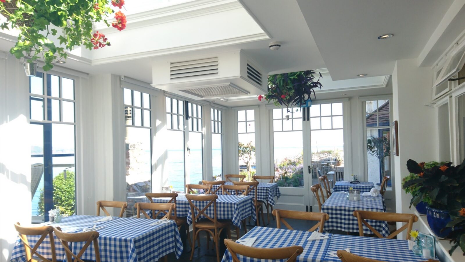 Sunlit restaurant with blue checkered tablecloths, wooden chairs, and large windows overlooking a serene outdoor view akin to the tranquil charm of Cary Arms, complete with suites and cozy beach huts nearby.