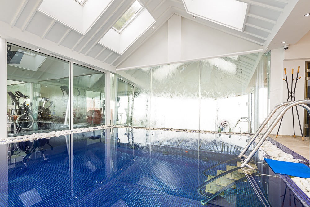 The Suites' indoor pool area features blue tiles, glass walls, exercise equipment, and a skylight ceiling. Whether you're swimming or unwinding after exploring the Cary Arms Beach Huts, this serene setting promises relaxation at its finest.