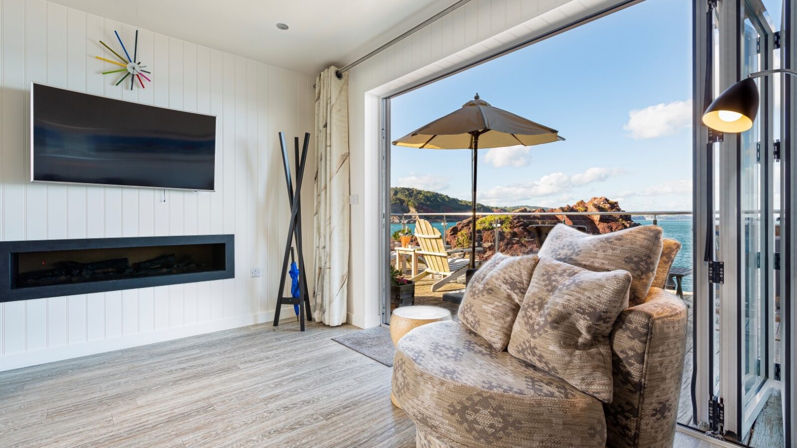 The cozy living room features a circular sofa and wall-mounted TV, with open doors leading to a seaside view reminiscent of charming beach huts at the Cary Arms. Enjoy outdoor seating that perfectly complements those tranquil suites by the sea.