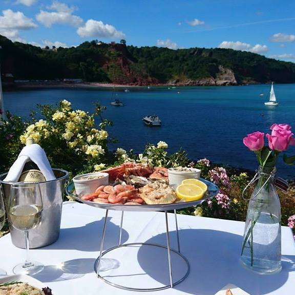 A seafood platter graces the table by the ocean at Cary Arms, with wine and pink flowers. Beyond, beach huts dot the coastline under a blue sky, as boats drift lazily.