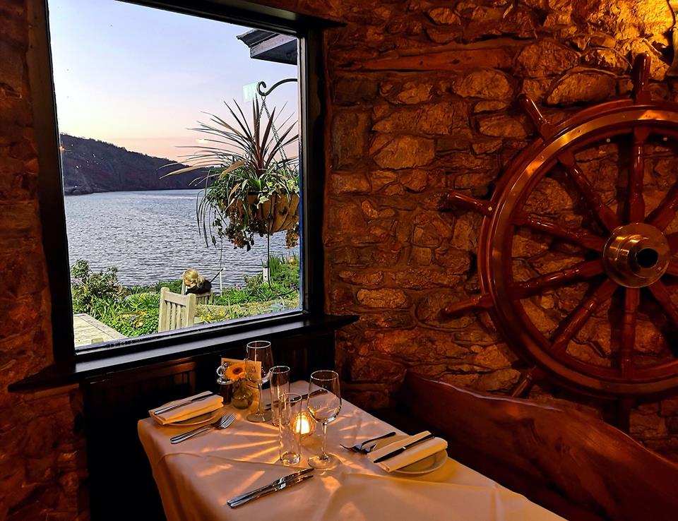 Cozy dining setting with a nautical theme, featuring a wooden ship wheel on the wall and a window view of the lake and sunset, reminiscent of the charming atmosphere at Cary Arms suites.