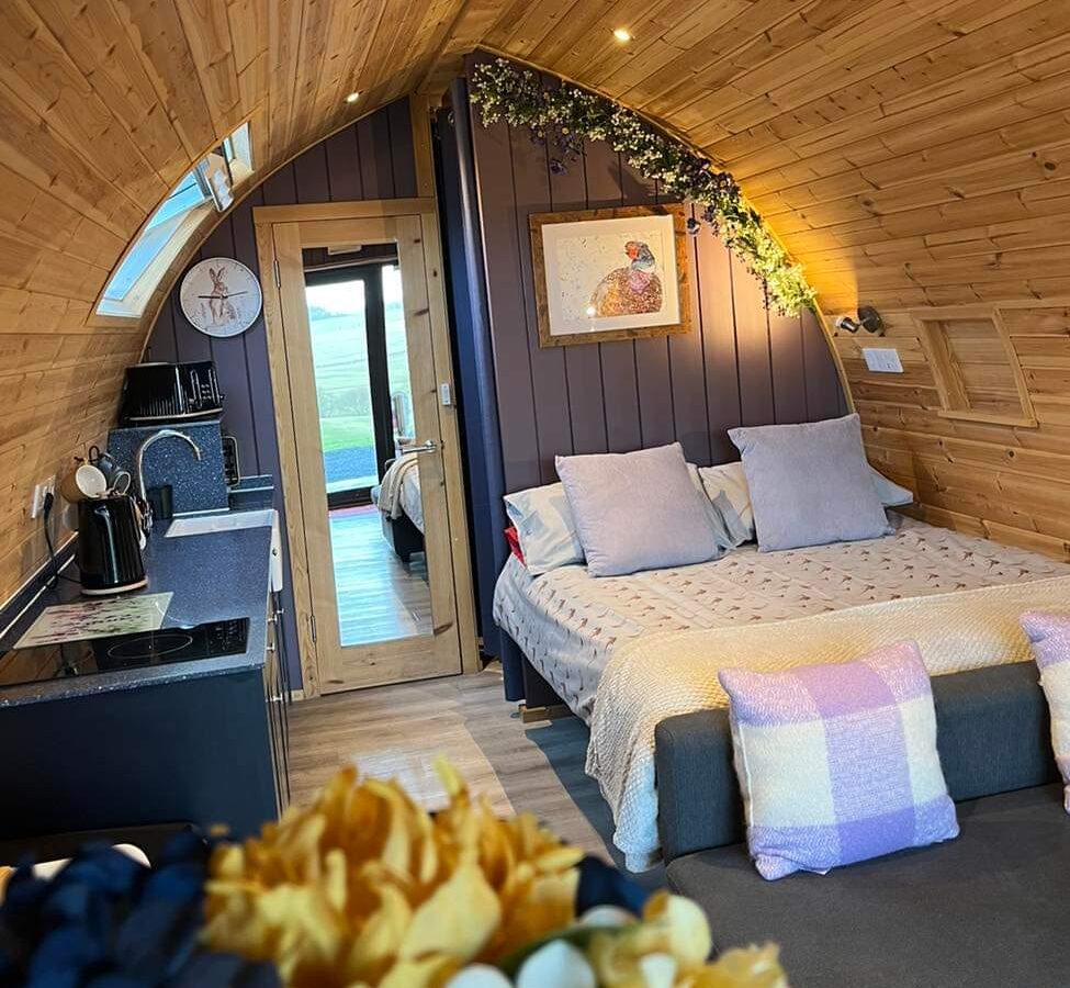 A cozy wooden cabin interior with a curved ceiling, featuring a neatly made bed adorned with pillows and blankets. To the left, there is a small kitchenette with a black countertop. The back door opens to a scenic view of Castle Guards Farm Retreat's lush outdoors. Flowers are in the foreground.