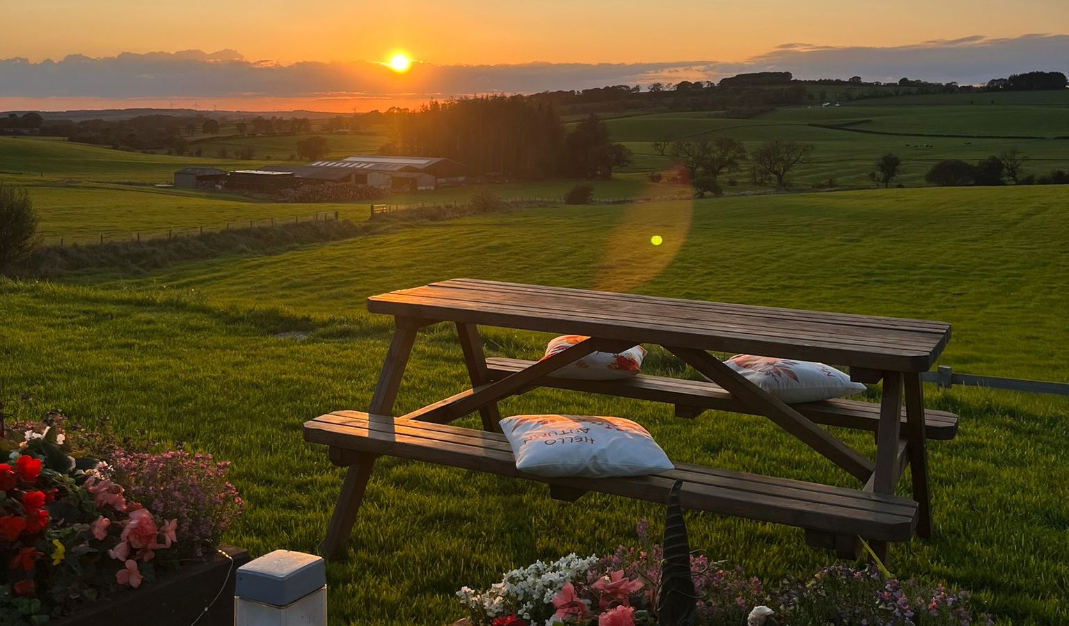 A wooden picnic table with two cushions is set on a lush green lawn, overlooking a scenic, expansive countryside. Beyond the table, the sun is setting, casting a warm glow over the rolling hills and distant trees. Vibrant flowers bloom in the garden of this charming farm retreat in the foreground.