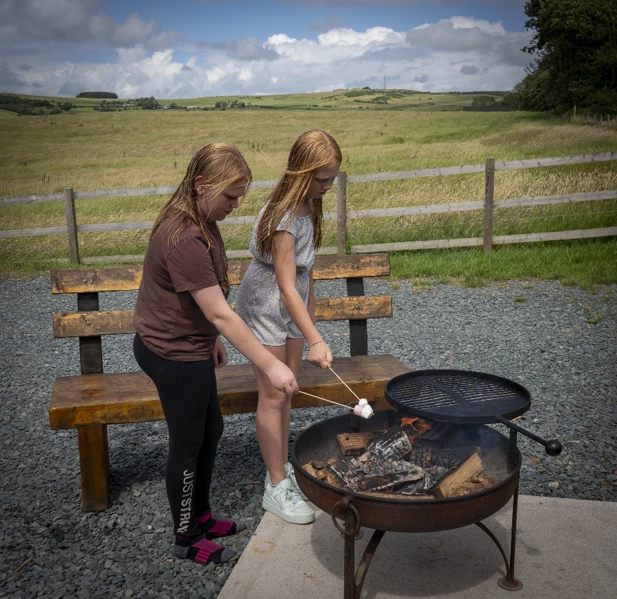 Two children roast marshmallows over a round fire pit on a sunny day at a cozy farm retreat. They stand on a gravel area in front of a wooden bench, with rolling fields and a clear, blue sky in the background. One child wears a brown shirt and black leggings; the other wears a light dress.