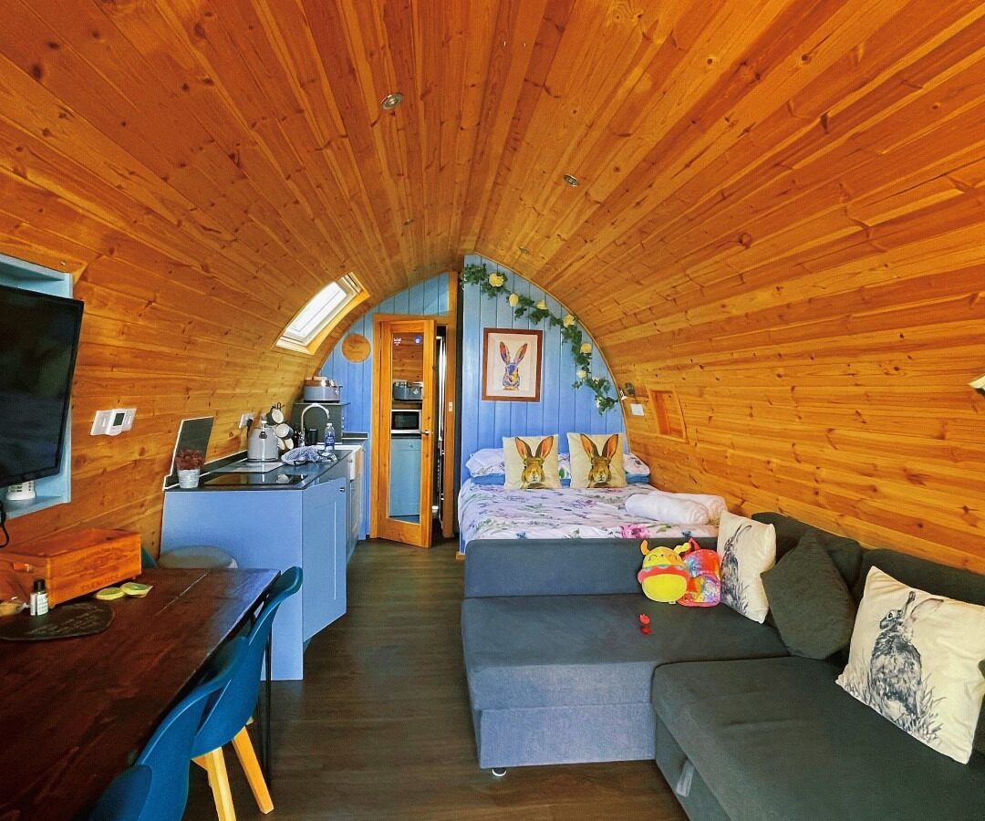 This image shows the interior of a cozy, wooden cabin with a curved ceiling. There's a seating area with a couch and pillows, a kitchen with blue cabinets, and a dining table with blue chairs. The back has a bed adorned with stuffed animals and floral decorations, perfect for a farm retreat.