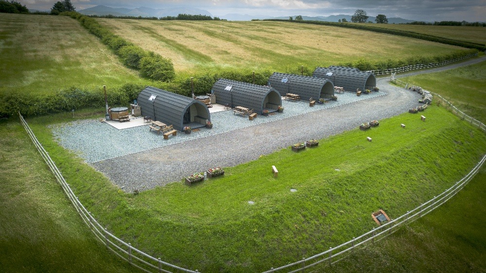 Aerial view of a scenic farm retreat with four curved, cabin-like structures lined up along a gravel pathway. Each cabin has a small seating area outside. The site is surrounded by expansive green fields and rolling hills, with a few trees visible in the background.