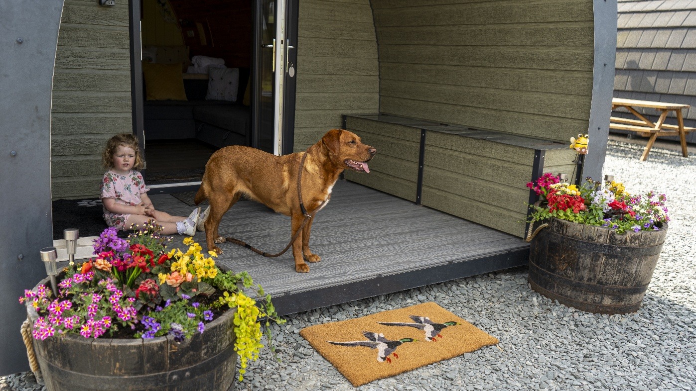 A small child and a brown dog sit by the entrance of a cozy wooden cabin at Castle Guards Farm, with colorful flowers in barrels on either side. A mat with bird designs lies on the gravel ground in front. Inside the cabin, a yellow cushion and green blanket are visible on a bench.