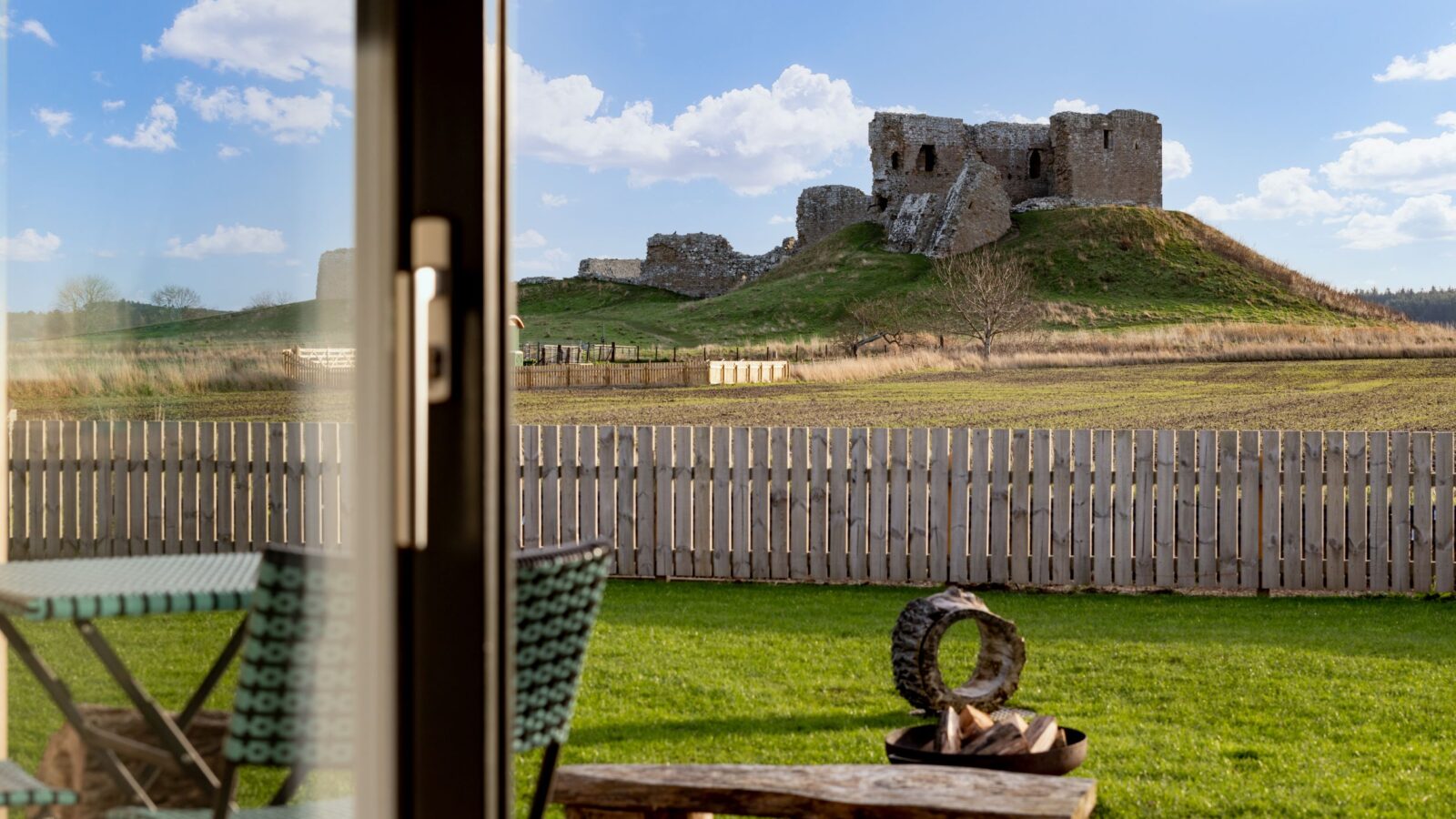 Seen through a window, a castle ruin perches on a grassy hill, framed by a wooden bench and fence in the foreground, evoking the rustic charm of a secluded cabin.