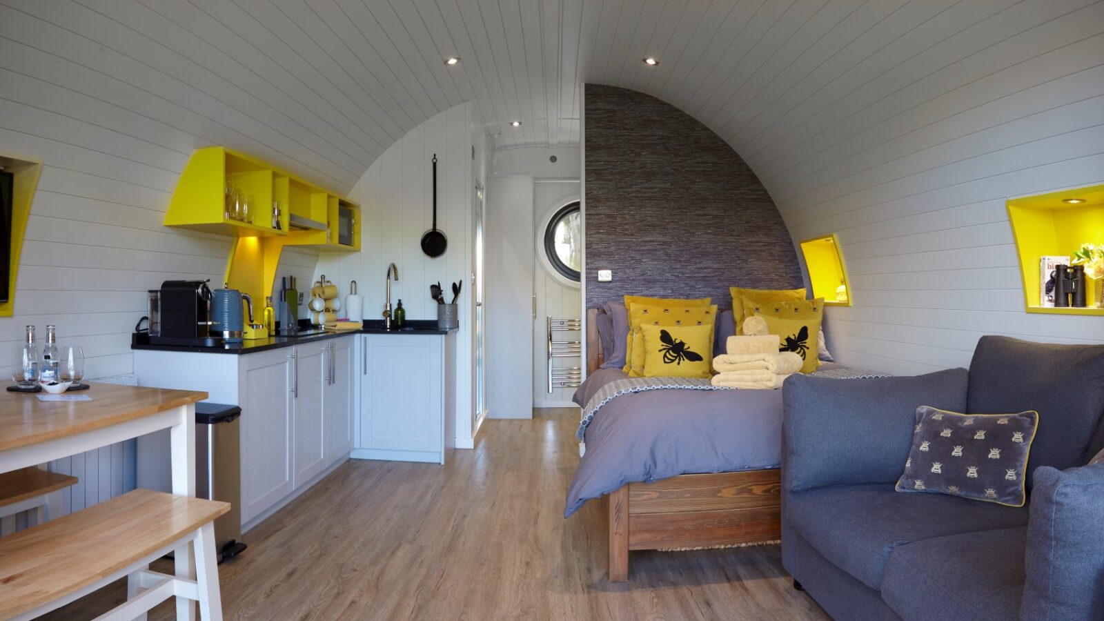 The cozy tiny home interior at Catgill Farm features a curved wooden ceiling, offering a rustic charm. Inside, you'll find a small kitchen with yellow shelves, a bed with yellow and gray accents, and a seating area. A round window frames the serene farmstay views beyond.