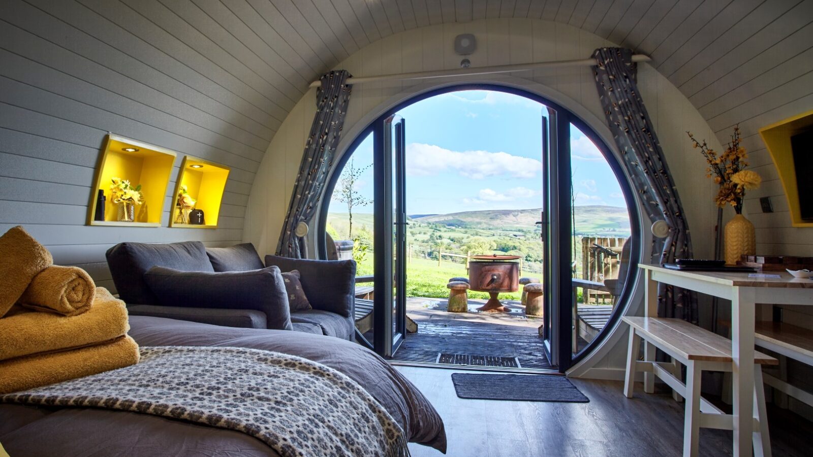 Cozy, modern cabin interior with a large circular glass door opening to a scenic view of the rolling hills and blue skies of the Yorkshire Dales. The room features a bed, couch, and small table with decorative items, creating a warm and inviting atmosphere at Catgill Farm.