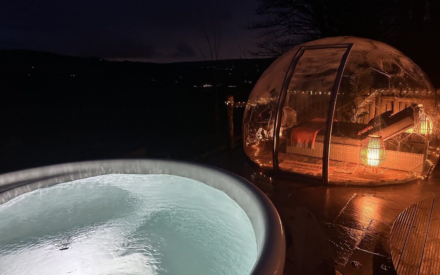 A nighttime scene at Catgill Farm features a glowing hot tub next to a cozy, illuminated bubble tent. Inside, the bed is adorned with warm lights, while the dark Yorkshire Dales provide a subtle outline of distant hills, perfect for an unforgettable farmstay experience.