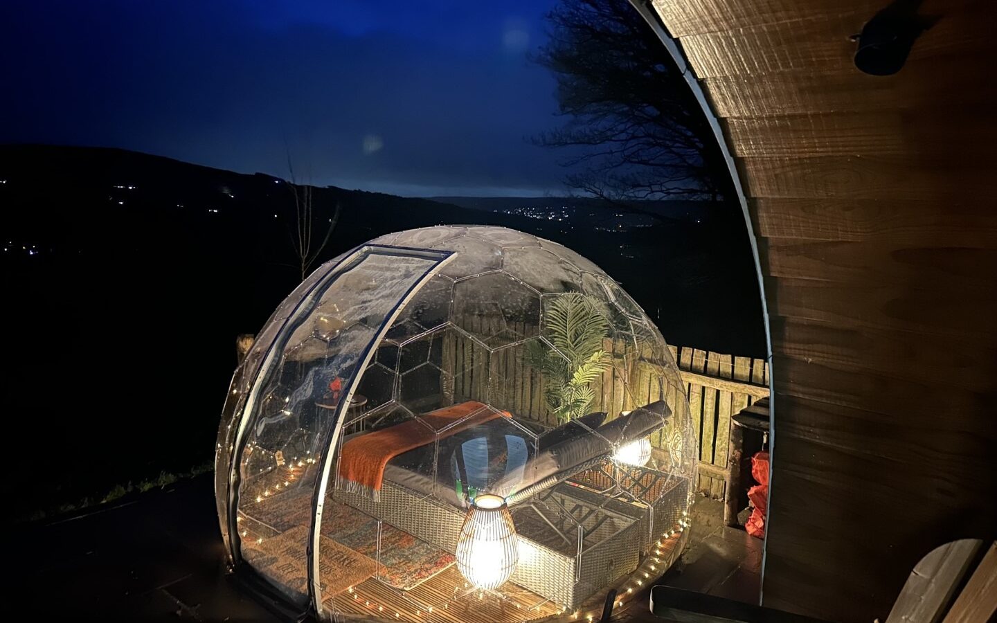 A transparent bubble tent at night, warmly lit with cozy seating and blankets, sits on a wooden deck overlooking a valley. Distant lights and trees stand against the dark sky, offering a serene countryside retreat at Catgill Farm.