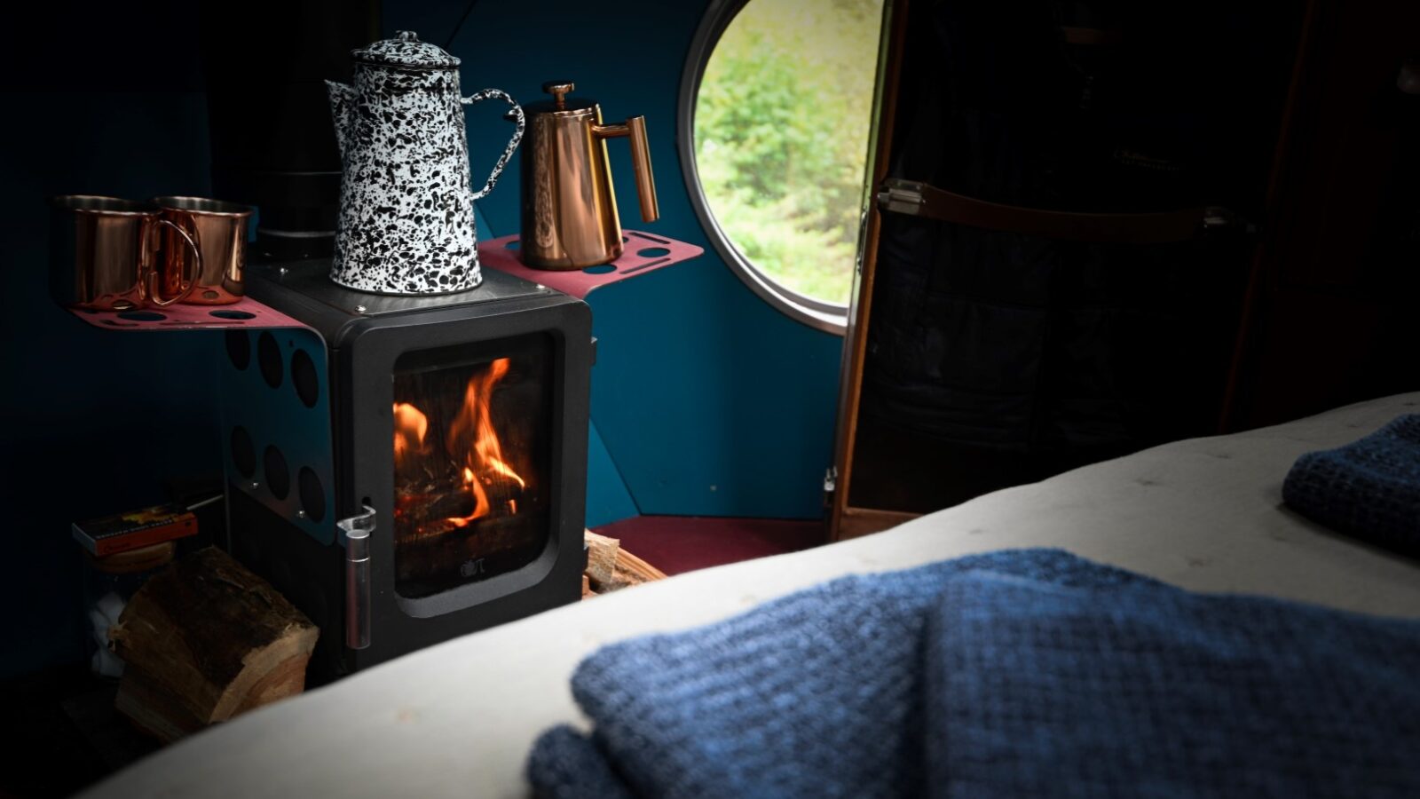 A cozy interior scene at Red Kite Glamping features a small lit wood stove with a black and white speckled kettle, a copper pot, and two copper mugs on it. Logs are stacked nearby. A round window offers a view of greenery outside, while folded blue blankets rest in the foreground on a bed in this Chillderness haven.