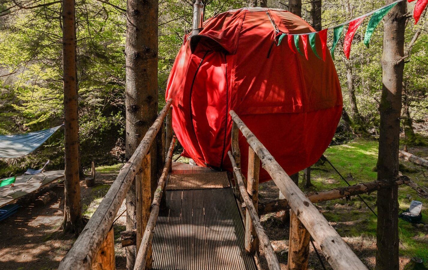 A spherical red tent, resembling a Red Kite, is suspended among trees in a forest. Accessible by a wooden footbridge, it's the epitome of glamping. Colorful pennant flags strung across the area add a festive touch to the lush, green Chillderness setting.