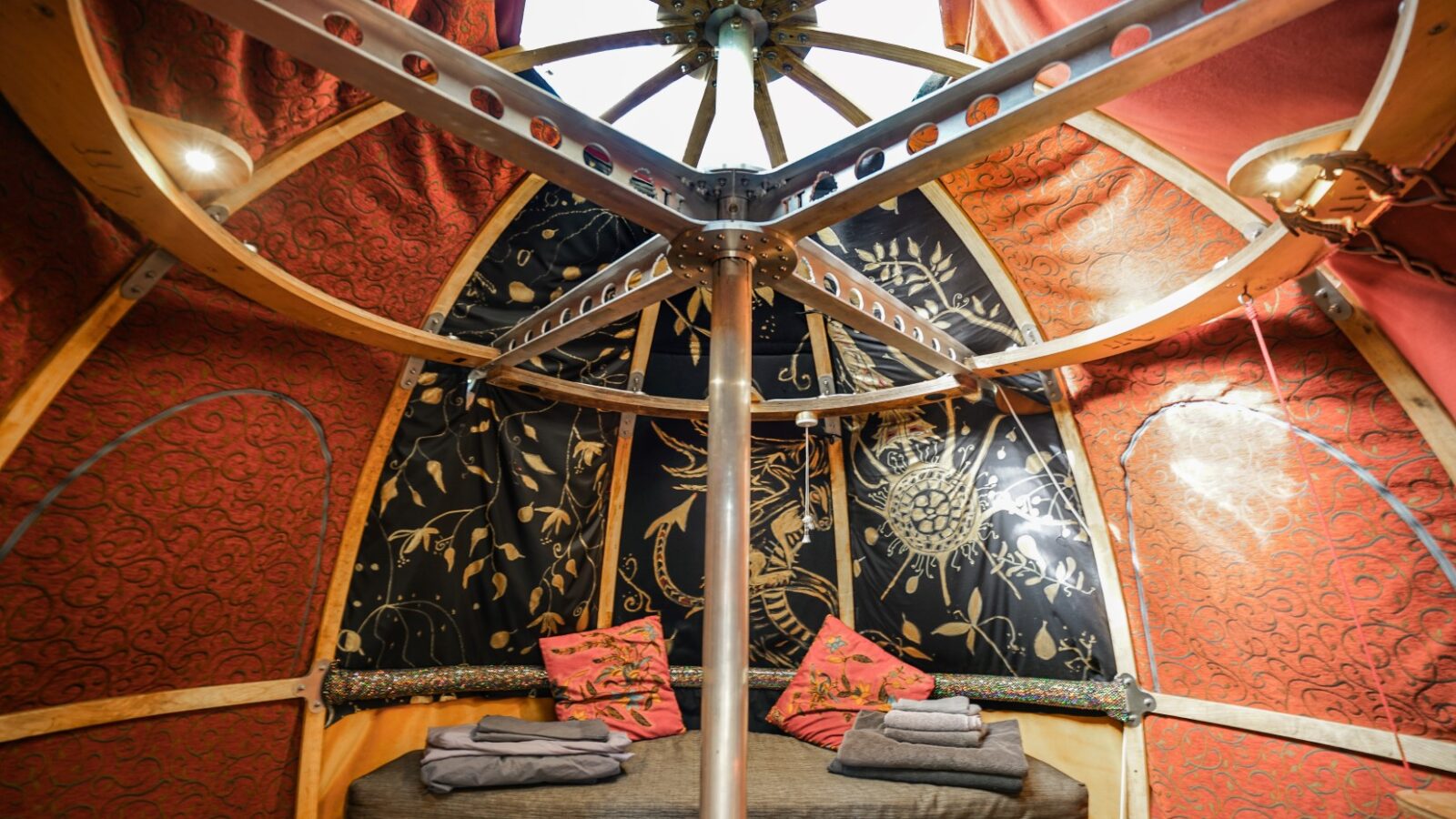 The image shows the interior of a well-decorated yurt, perfect for glamping, with intricate patterns on the fabric walls and ceiling. A metal pole supports the structure. There is a cushioned seating area adorned with decorative red and pink pillows, and folded towels are placed on the seat—a true Chillderness retreat.