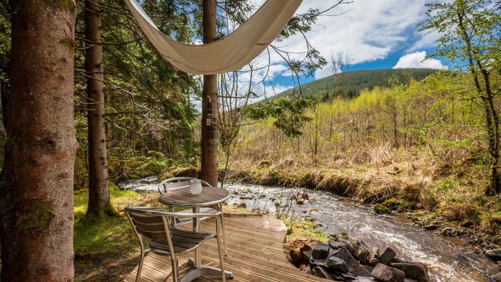 A small wooden deck with two chairs and a table set for one is nestled among tall trees by a serene creek. A hammock is strung between the trees, offering a peaceful view of the lush, green landscape—a perfect glamping spot with sightings of red kites soaring against a partly cloudy sky.