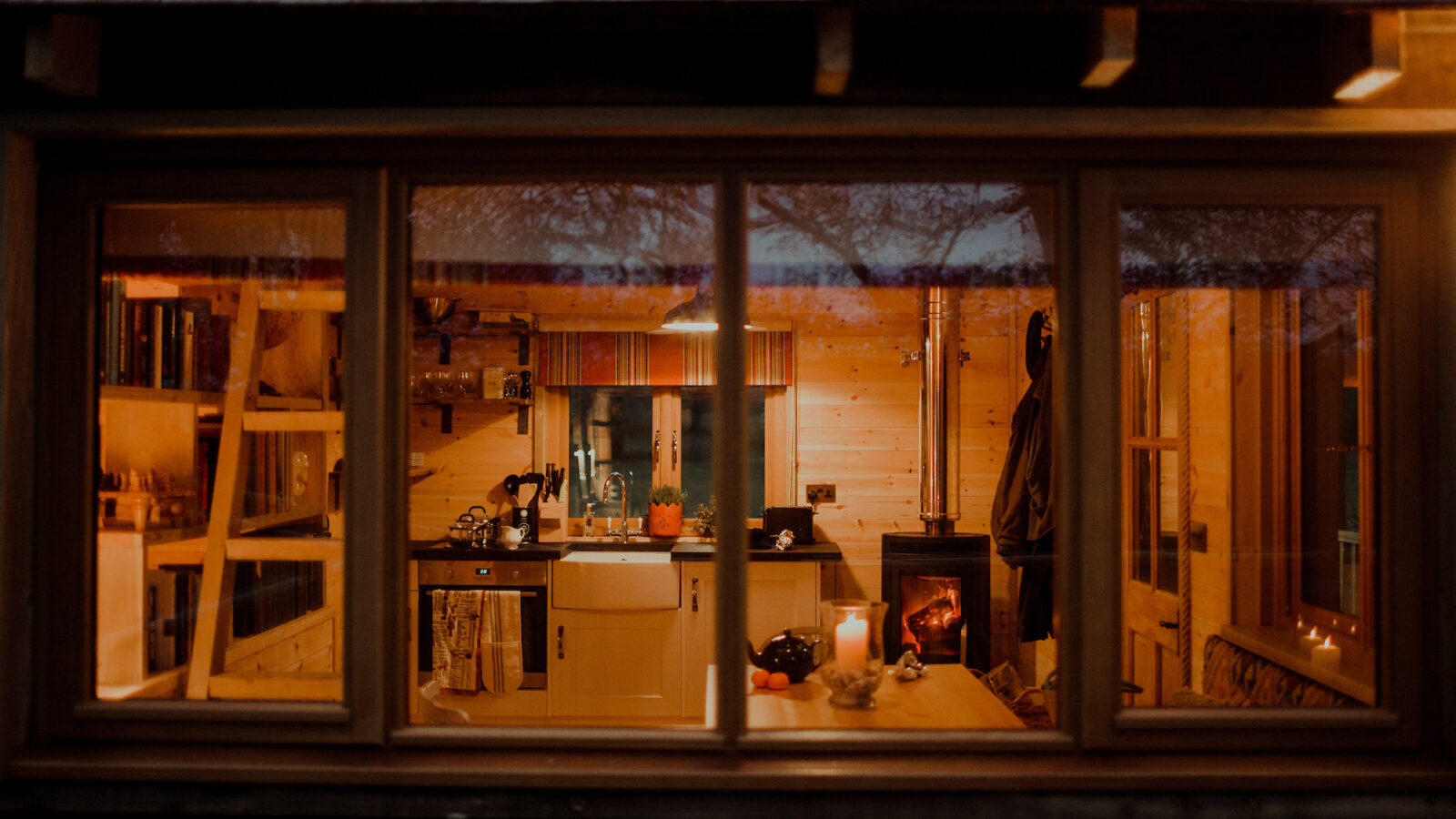 A cozy cabin kitchen and dining area are warmly lit by candlelight and a wood-burning stove. The room, reminiscent of the Netherby Treehouse, features wooden walls, a farmhouse sink, a ladder leading to a loft, and shelves stocked with various items. The view is from outside through a large window.