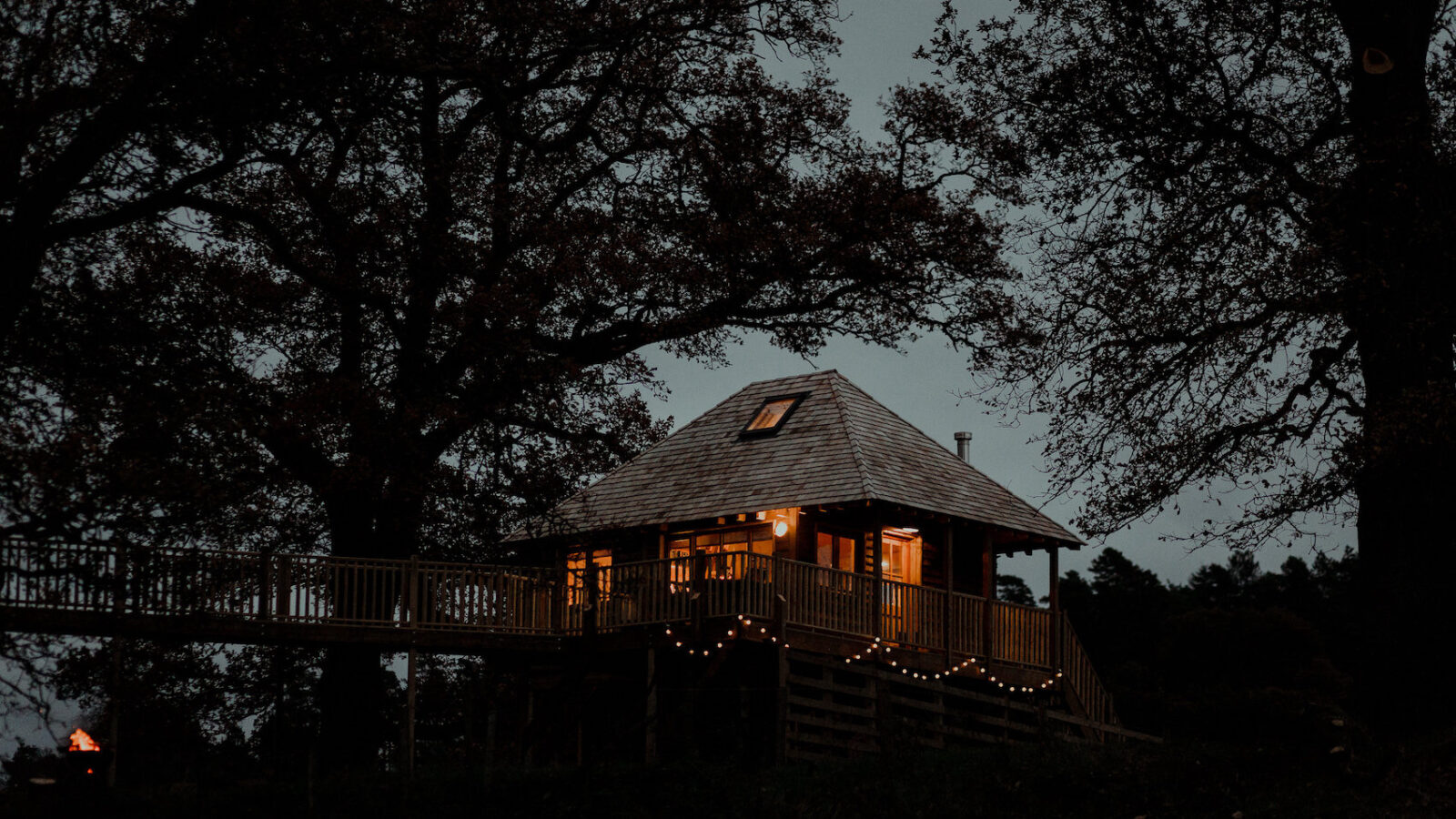 A cozy cabin illuminated with warm lights sits nestled among large, dark trees in an evening setting, reminiscent of the Netherby Treehouse. The sky is dusky, and soft lights strung along the railings add a gentle glow. The atmosphere feels serene and inviting.