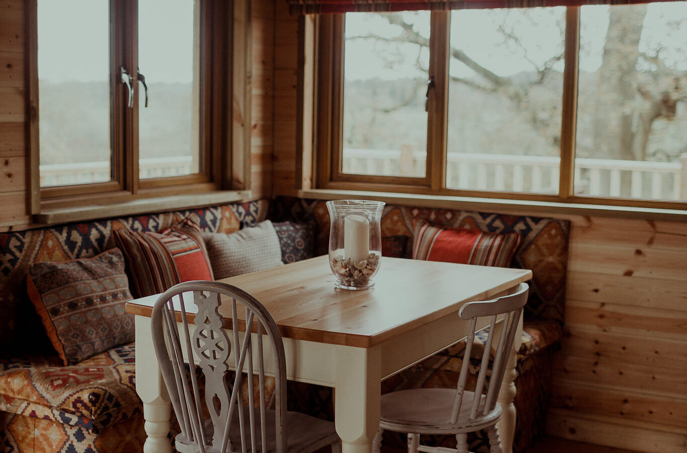 A cozy wood-paneled dining area at Netherby Treehouse features a square wooden table with two white chairs. A bench with patterned cushions lines the wall under two windows. The space is adorned with warm, earth-toned rugs and a clear glass vase sits atop the table.