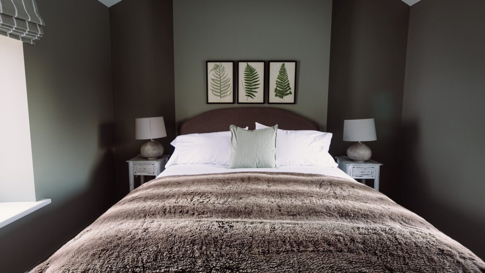 A bedroom with dark gray walls features a bed with white bedding, a gray throw blanket, and a large white pillow. Two bedside tables, each with a lamp, flank the bed. Above the bed are three framed botanical prints reminiscent of Cleugh Foot. A window with a striped Roman shade is on the left.