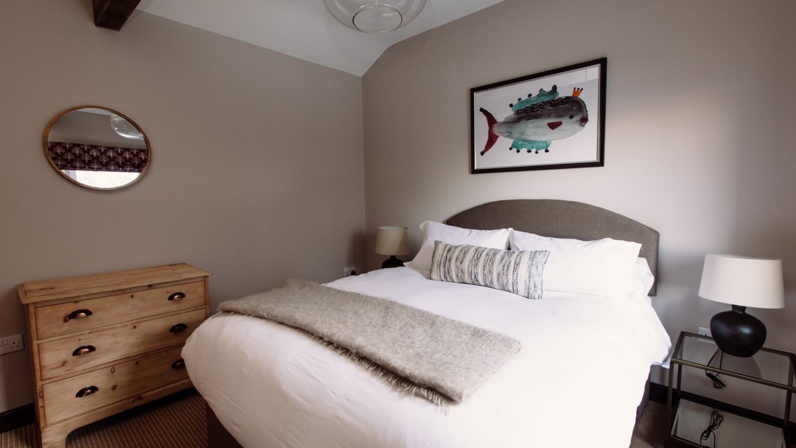 A cozy bedroom at Cleugh Foot features a neatly made bed with white linens and grey accents. A colorful fish painting hangs above the bed. The room includes a wooden dresser with a circular mirror above it and two bedside tables with lamps. A wooden beam graces the ceiling.