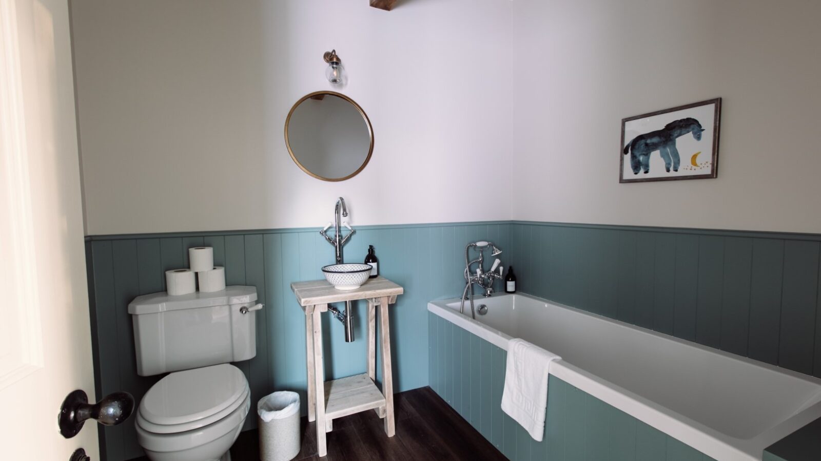 A Cleugh Foot bathroom features a toilet, a small sink on a wooden stand, and a bathtub with a towel hanging over the edge. Above the sink is a round mirror, and above the bathtub is a framed picture of a blue horse. The walls are painted in light grey and teal.