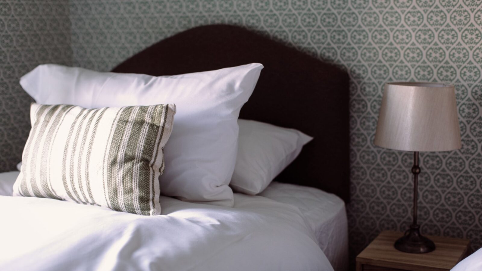 A neatly made bed with white pillows and a striped cushion stands next to a wooden nightstand and a beige lamp. The wall behind the bed features a green and white patterned wallpaper, reminiscent of Cleugh Foot's charming interiors. The scene is well-lit, creating a cozy and inviting atmosphere.