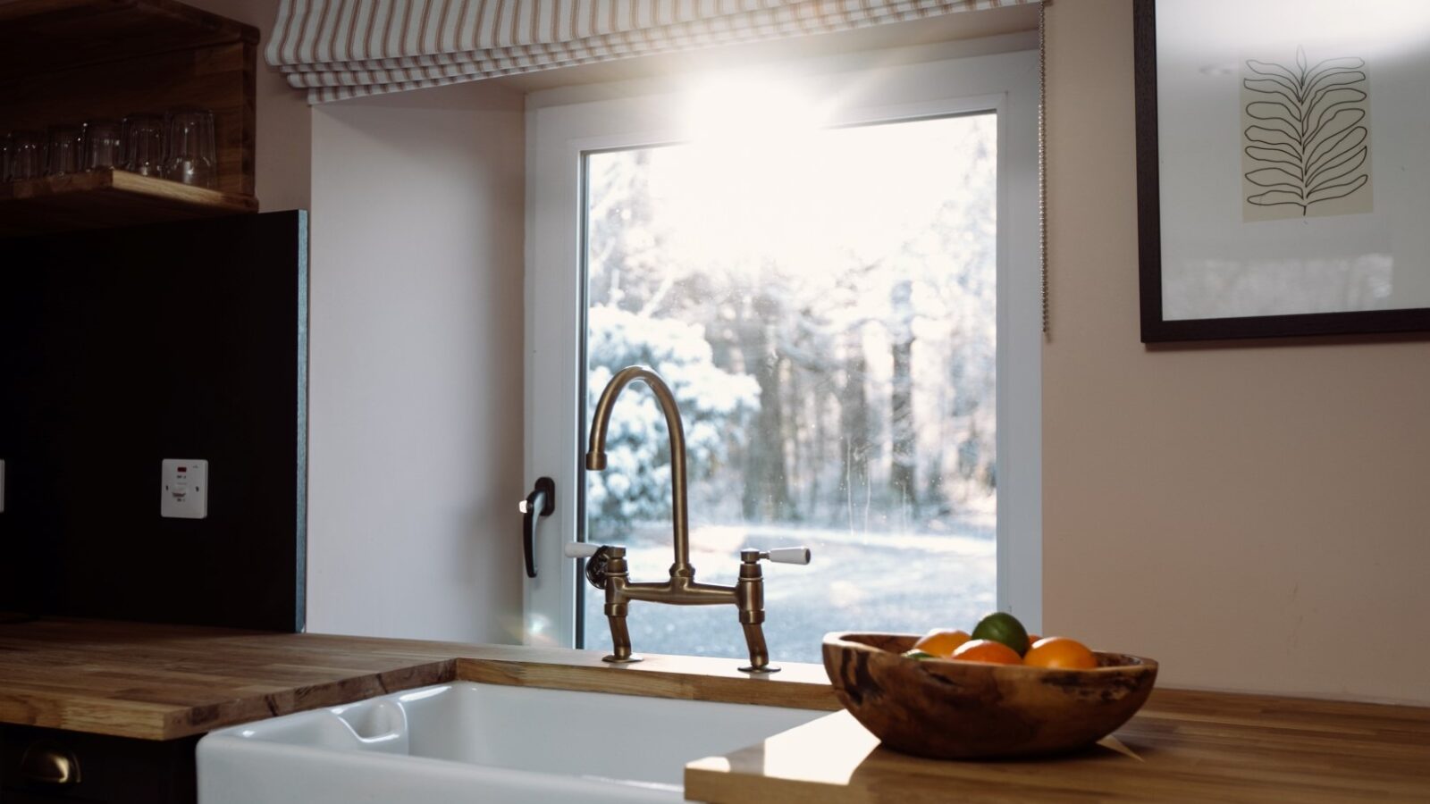 A cozy kitchen scene at Cleugh Foot features a farmhouse sink below a large window with light streaming through. A wooden bowl filled with fruit sits on the counter, and a piece of minimalist artwork hangs on the wall. A white and beige striped valance covers the top of the window.