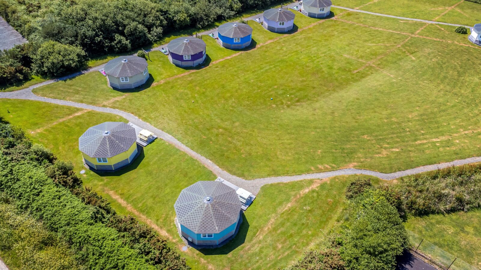 Aerial view of eight round, blue-roofed cabins evenly spaced on a large grassy area with intersecting pathways, evoking a coastal charm.