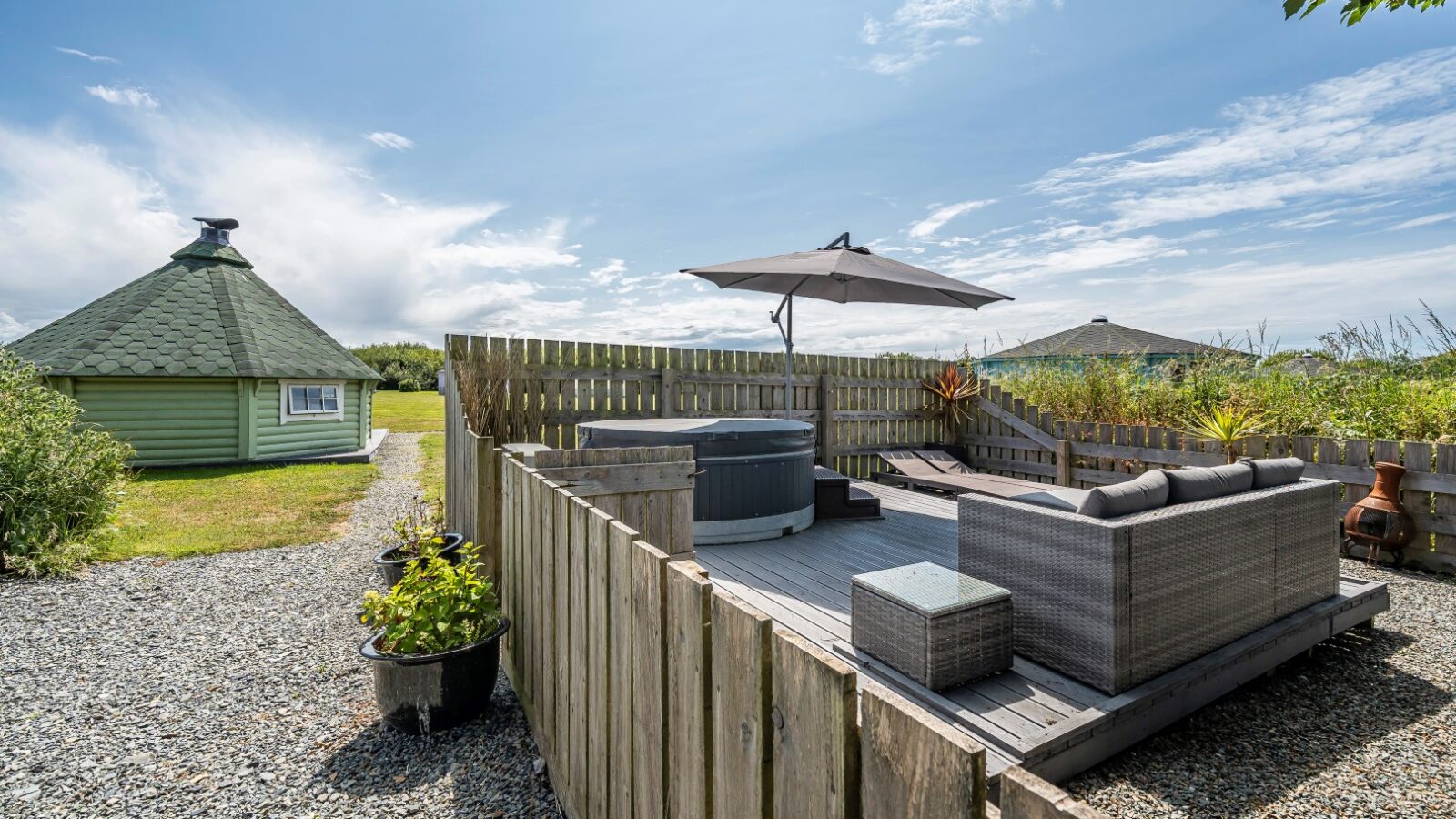 A cozy outdoor patio with a hot tub, rattan furniture, and an umbrella awaits you on a sunny day, nestled next to a charming green wooden cabin.