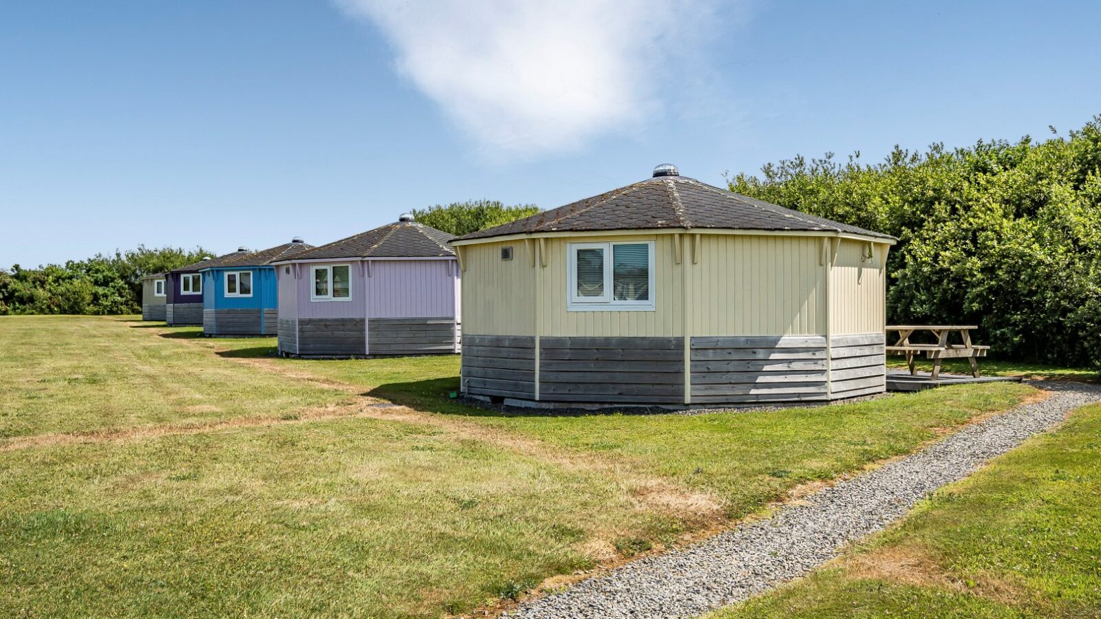 Four round coastal cabins, each painted in soothing pastel colors, stand on a grassy field under a clear blue sky, connected by a charming gravel path.