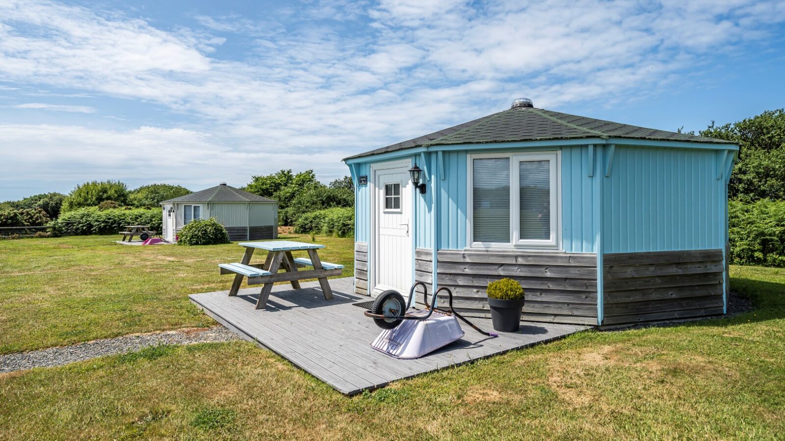 A cozy, light blue coastal cabin with a wooden deck and picnic table sits among garden tools in a grassy area under a partly cloudy sky.